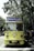 A vintage-style yellow bus with 'Bandung Tour on Bus' written on the front, parked on a tree-lined street. The bus has round headlights and a distinctive front design. Several people are standing nearby, some wearing masks. The surrounding environment is lush with green trees, and there's a streetlamp with a globe light.