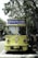 A vintage-style yellow bus with 'Bandung Tour on Bus' written on the front, parked on a tree-lined street. The bus has round headlights and a distinctive front design. Several people are standing nearby, some wearing masks. The surrounding environment is lush with green trees, and there's a streetlamp with a globe light.