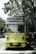 A vintage-style yellow bus with 'Bandung Tour on Bus' written on the front, parked on a tree-lined street. The bus has round headlights and a distinctive front design. Several people are standing nearby, some wearing masks. The surrounding environment is lush with green trees, and there's a streetlamp with a globe light.