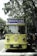 A vintage-style yellow bus with 'Bandung Tour on Bus' written on the front, parked on a tree-lined street. The bus has round headlights and a distinctive front design. Several people are standing nearby, some wearing masks. The surrounding environment is lush with green trees, and there's a streetlamp with a globe light.