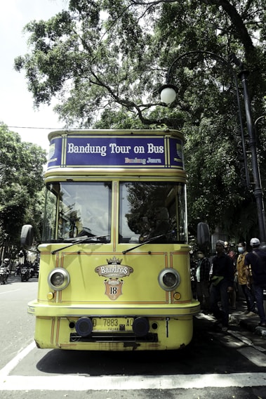 A vintage-style yellow bus with 'Bandung Tour on Bus' written on the front, parked on a tree-lined street. The bus has round headlights and a distinctive front design. Several people are standing nearby, some wearing masks. The surrounding environment is lush with green trees, and there's a streetlamp with a globe light.