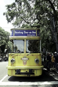 A vintage-style yellow bus with 'Bandung Tour on Bus' written on the front, parked on a tree-lined street. The bus has round headlights and a distinctive front design. Several people are standing nearby, some wearing masks. The surrounding environment is lush with green trees, and there's a streetlamp with a globe light.