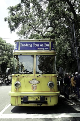 A vintage-style yellow bus with 'Bandung Tour on Bus' written on the front, parked on a tree-lined street. The bus has round headlights and a distinctive front design. Several people are standing nearby, some wearing masks. The surrounding environment is lush with green trees, and there's a streetlamp with a globe light.