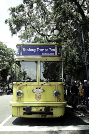 A vintage-style yellow bus with 'Bandung Tour on Bus' written on the front, parked on a tree-lined street. The bus has round headlights and a distinctive front design. Several people are standing nearby, some wearing masks. The surrounding environment is lush with green trees, and there's a streetlamp with a globe light.