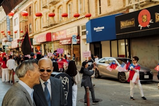 A busy street scene features people participating in a parade or rally, holding various flags including Taiwanese and American flags. The area is adorned with red lanterns and various store signs. Two older men in suits are conversing in the foreground, while others take photos and interact in the background.