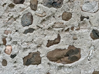 A textured surface featuring a mix of gray cement and embedded rocks of varying sizes and colors. The rocks appear naturally weathered and are unevenly distributed, creating an irregular pattern.
