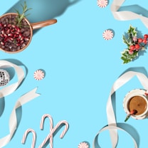 A festive holiday flat lay arranged on a bright blue background, featuring a cup of cocoa with candy cane stirrers, a bowl with pomegranate seeds and rosemary, peppermint candies, candy canes, holly with red berries, a white ribbon, and a small disco ball ornament.