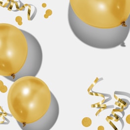 Several shiny golden balloons float alongside curling ribbons and scattered confetti on a light background, casting soft shadows.