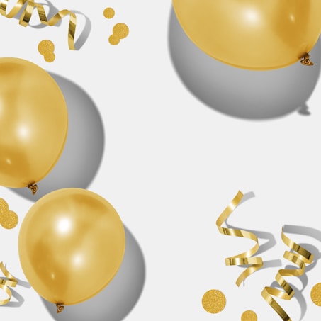 Several shiny golden balloons float alongside curling ribbons and scattered confetti on a light background, casting soft shadows.