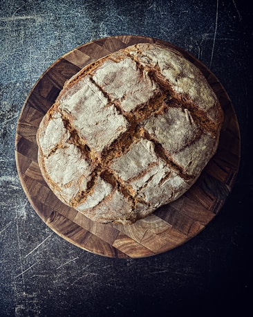 A rustic loaf of bread with a crusty exterior sits on a wooden cutting board. The bread has a golden brown color and is marked with slashes on the top, creating a decorative pattern. The background is a textured dark surface, enhancing the warmth and homely feel of the bread.