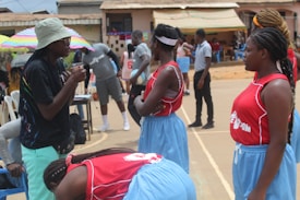 A group of young women in red and blue sports uniforms stand on a basketball court, appearing to listen to a coach or teammate. There are people and colorful umbrellas in the background, indicating an outdoor setting with possibly warm weather.