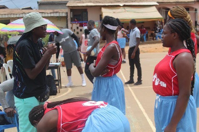 A group of young women in red and blue sports uniforms stand on a basketball court, appearing to listen to a coach or teammate. There are people and colorful umbrellas in the background, indicating an outdoor setting with possibly warm weather.
