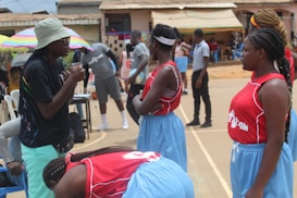 A group of young women in red and blue sports uniforms stand on a basketball court, appearing to listen to a coach or teammate. There are people and colorful umbrellas in the background, indicating an outdoor setting with possibly warm weather.