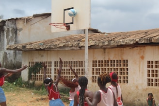 A group of young girls wearing red jerseys and blue shorts are actively engaged in playing basketball. The scene takes place outdoors against a backdrop of a weathered building with a tin roof. A basketball is in mid-air, about to go through the hoop, while the players eagerly reach upwards with their arms extended.