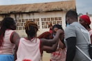 A group of young athletes wearing uniforms, gathered in a close circle, appear to be engaged in a team huddle or pep talk. The setting is outdoors, in front of an old building with a rusted roof. One person is wearing a shirt labeled 'COACH', suggesting they are giving instructions or encouragement. The mood seems supportive and focused.
