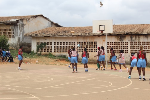 A group of young women are playing a basketball game on an outdoor court. They are wearing light blue shorts and red or white jerseys, with some spectators seated on the side. The court is surrounded by old buildings with damaged roofs and worn walls.