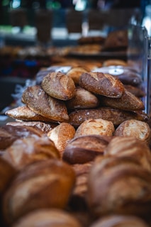A variety of artisan breads are displayed in a bakery setting. The loaves have a rustic appearance with golden-brown crusts, some dusted with flour. They are arranged in a pyramid shape, creating a visually appealing display under soft lighting.