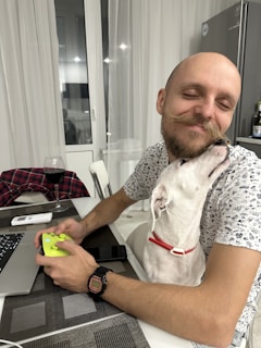 A man is sitting at a table, holding a gaming controller and smiling with his eyes closed as a white dog with a red collar affectionately nuzzles his face. On the table, there's a laptop, a glass of red wine, a smartphone, and a folded flannel shirt.