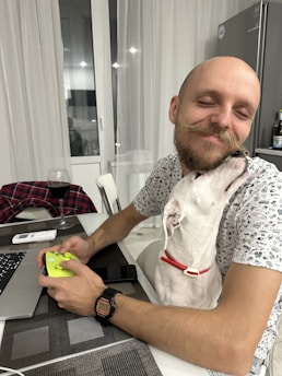A man is sitting at a table, holding a gaming controller and smiling with his eyes closed as a white dog with a red collar affectionately nuzzles his face. On the table, there's a laptop, a glass of red wine, a smartphone, and a folded flannel shirt.