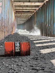 An industrial corridor made of rusted metal beams creates a tunnel-like structure along a rough, pebbly shoreline. Water splashes against the sides due to waves pushing in from one end. A weathered, orange oil drum lies on the rocky ground, with visible text indicating it once contained oil from Japan.