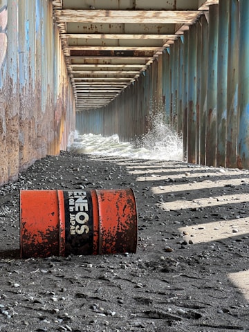 An industrial corridor made of rusted metal beams creates a tunnel-like structure along a rough, pebbly shoreline. Water splashes against the sides due to waves pushing in from one end. A weathered, orange oil drum lies on the rocky ground, with visible text indicating it once contained oil from Japan.