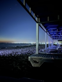 A nighttime beach scene with several empty lounge chairs under a pergola. The beach is illuminated by distant city lights reflecting across the water. The sky is clear, with a gradient from a dark blue to a lighter hue near the horizon.