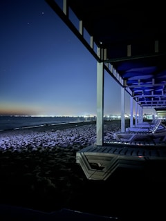 A nighttime beach scene with several empty lounge chairs under a pergola. The beach is illuminated by distant city lights reflecting across the water. The sky is clear, with a gradient from a dark blue to a lighter hue near the horizon.