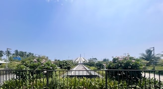 A well-maintained walkway surrounded by lush greenery, with blooming flowers and tall palm trees. In the background, a large dome-shaped structure is visible under a clear blue sky.