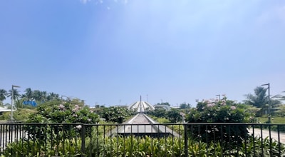 A well-maintained walkway surrounded by lush greenery, with blooming flowers and tall palm trees. In the background, a large dome-shaped structure is visible under a clear blue sky.