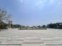 A wide plaza with a patterned stone pavement, surrounded by green hedges and palm trees. In the center, there is a modern architectural structure with people walking around it. The sky is clear and blue, creating a tranquil atmosphere.
