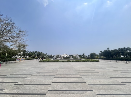 A wide plaza with a patterned stone pavement, surrounded by green hedges and palm trees. In the center, there is a modern architectural structure with people walking around it. The sky is clear and blue, creating a tranquil atmosphere.
