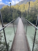 A narrow suspension bridge over a rocky mountain creek surrounded by green forest