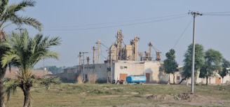 An industrial building with large machinery in a rural area, surrounded by trees and power lines. The setting includes a few palm trees and a grassy foreground with a blue water tanker parked beside the structure.