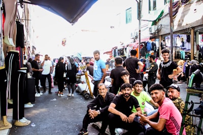 A busy market street with a group of people gathered in the foreground, some sitting on bags and others standing. Shops line the street, each displaying various clothing items. Mannequins dressed in pants are visible on the left. The atmosphere appears lively and crowded, with many people interacting and walking around.
