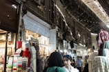 A bustling marketplace scene with various textiles and fabrics displayed on stands. Several people are walking through the narrow aisles beneath an arched, metal-roofed structure. The lighting is a mix of natural and artificial, casting shadows and highlights across the scene.