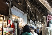 A bustling marketplace scene with various textiles and fabrics displayed on stands. Several people are walking through the narrow aisles beneath an arched, metal-roofed structure. The lighting is a mix of natural and artificial, casting shadows and highlights across the scene.