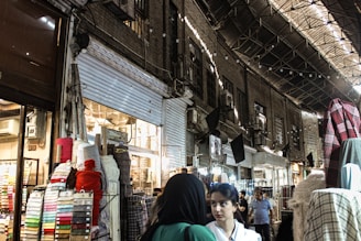 A bustling marketplace scene with various textiles and fabrics displayed on stands. Several people are walking through the narrow aisles beneath an arched, metal-roofed structure. The lighting is a mix of natural and artificial, casting shadows and highlights across the scene.