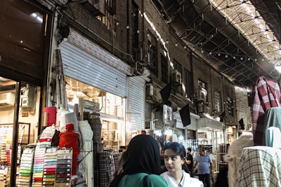A bustling marketplace scene with various textiles and fabrics displayed on stands. Several people are walking through the narrow aisles beneath an arched, metal-roofed structure. The lighting is a mix of natural and artificial, casting shadows and highlights across the scene.