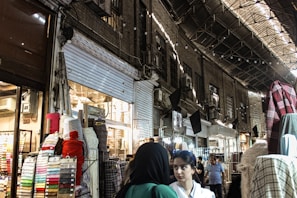 A bustling marketplace scene with various textiles and fabrics displayed on stands. Several people are walking through the narrow aisles beneath an arched, metal-roofed structure. The lighting is a mix of natural and artificial, casting shadows and highlights across the scene.
