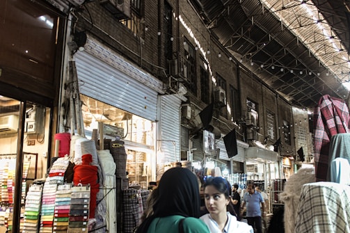 A bustling marketplace scene with various textiles and fabrics displayed on stands. Several people are walking through the narrow aisles beneath an arched, metal-roofed structure. The lighting is a mix of natural and artificial, casting shadows and highlights across the scene.