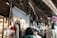 A bustling marketplace scene with various textiles and fabrics displayed on stands. Several people are walking through the narrow aisles beneath an arched, metal-roofed structure. The lighting is a mix of natural and artificial, casting shadows and highlights across the scene.