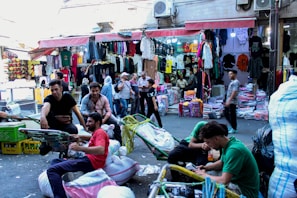 A bustling street market scene with several people engaged in various activities. There are vendors displaying a variety of colorful clothes hanging in stalls. Shoppers, some carrying bags, move through the market. A few men are sitting on trolleys or near stacks of merchandise, possibly taking a break or preparing goods for transport.