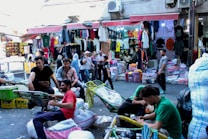 A bustling street market scene with several people engaged in various activities. There are vendors displaying a variety of colorful clothes hanging in stalls. Shoppers, some carrying bags, move through the market. A few men are sitting on trolleys or near stacks of merchandise, possibly taking a break or preparing goods for transport.