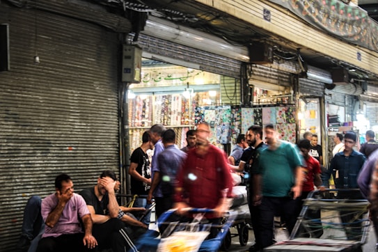 A bustling market scene with people walking, some carrying goods. Shops in the background have colorful displays and signs. The overall atmosphere is lively and crowded, with a mix of people engaged in different activities.