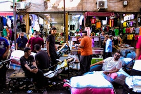 A bustling marketplace scene with people engaging in various activities. Stalls are set up displaying colorful clothing, and shoppers are interacting with vendors. Several men are seated on a cart filled with bags, while others are standing or walking around. The environment is lively and crowded with an array of merchandise visible in the background.