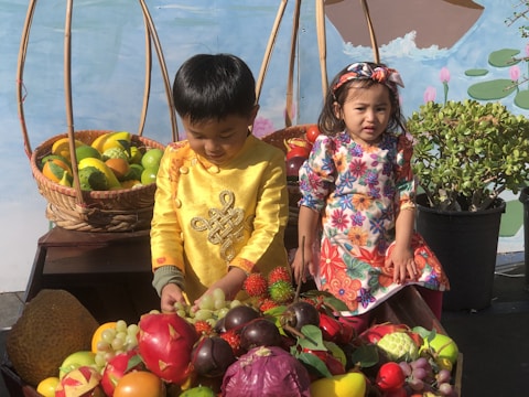 Two children wearing colorful traditional clothing are surrounded by a variety of fruits and vegetables in wicker baskets. One child is sitting on a wooden surface while the other is standing beside a basket. The background features a painted mural with a water and plant design.