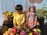 Two children wearing colorful traditional clothing are surrounded by a variety of fruits and vegetables in wicker baskets. One child is sitting on a wooden surface while the other is standing beside a basket. The background features a painted mural with a water and plant design.
