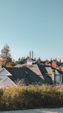 A residential area with a house featuring a dark roof and stone chimney, surrounded by greenery and trees displaying autumn colors. Two tall chimneys or towers are visible in the background, against a clear blue sky.