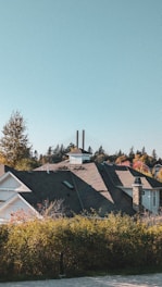 A residential area with a house featuring a dark roof and stone chimney, surrounded by greenery and trees displaying autumn colors. Two tall chimneys or towers are visible in the background, against a clear blue sky.