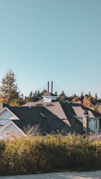 A residential area with a house featuring a dark roof and stone chimney, surrounded by greenery and trees displaying autumn colors. Two tall chimneys or towers are visible in the background, against a clear blue sky.